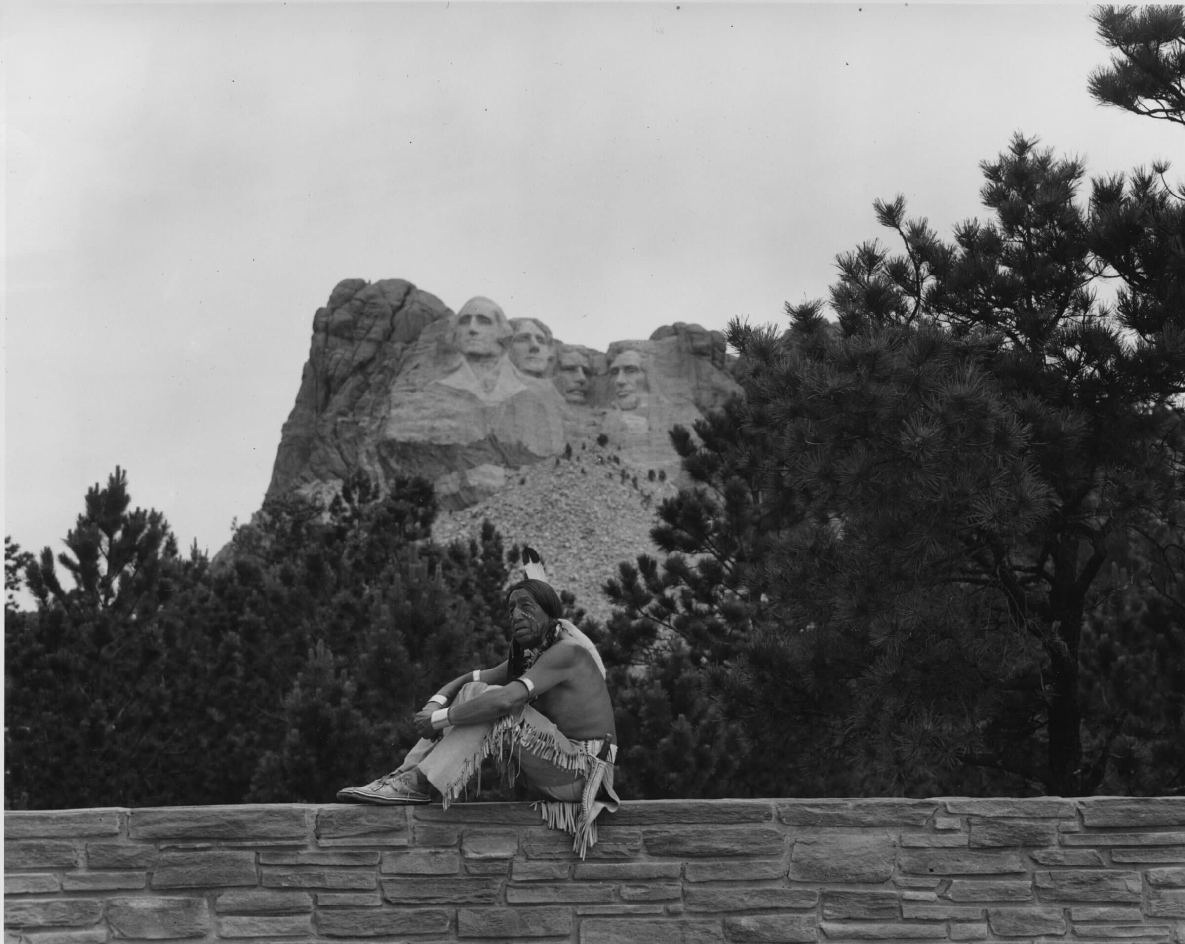 Native American man dressed in traditional clothing sitting on a stone wall at the base of Mount Rushmore; he has white dots on his face and braided hair.