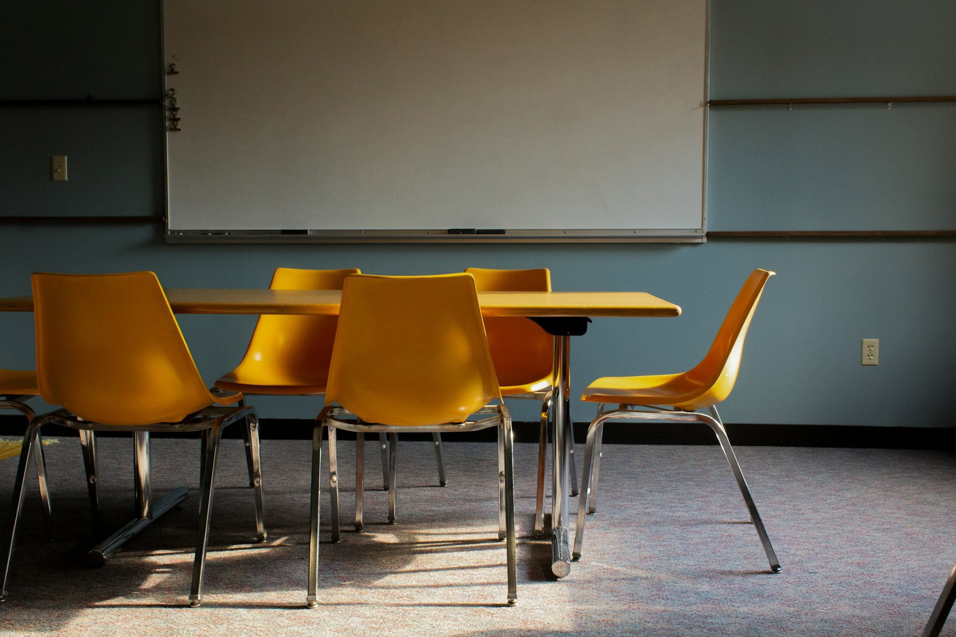 Five yellow chairs with metal legs around a yellow chair with metal legs in a classroom in front of a whiteboard on a blue wall.