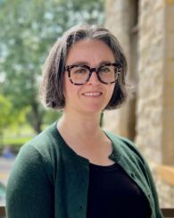 Smiling woman in green cardigan with dark rimmed glasses in front of trees and a brick building