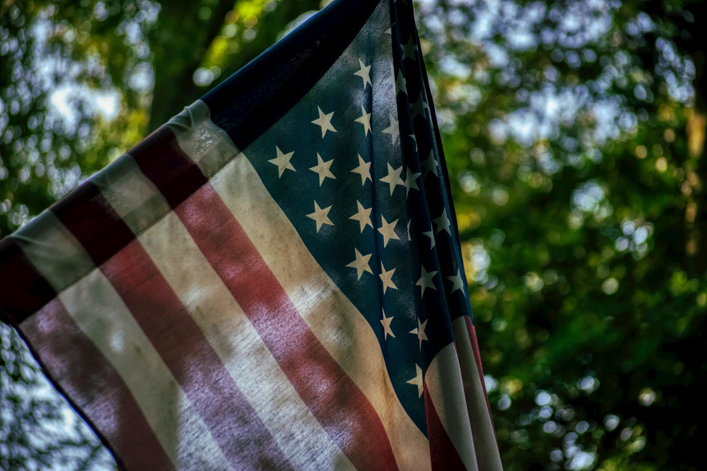 Faded and weathered American flag hanging on a diagonal pole with blurry trees in background.