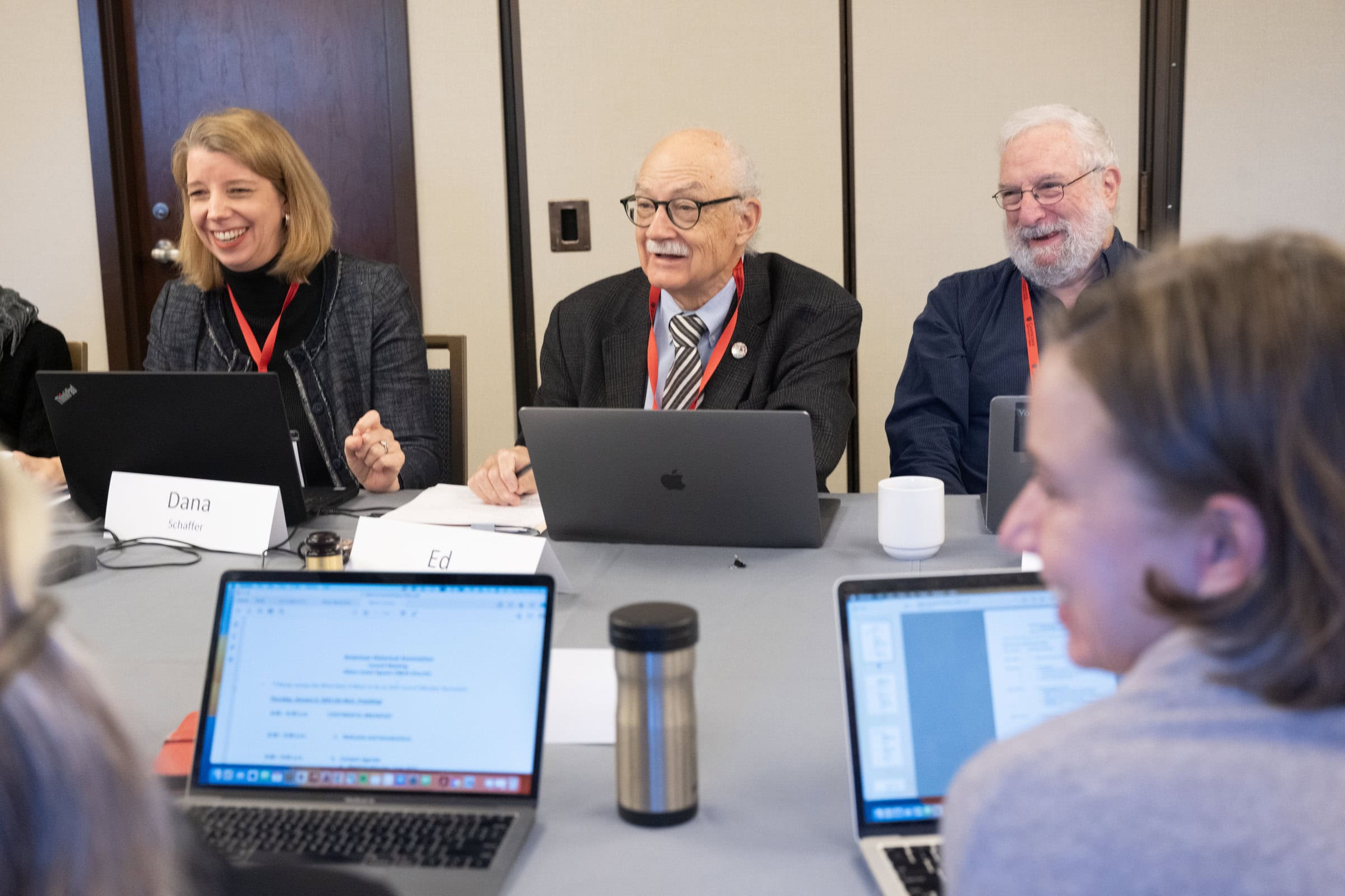 A blonde woman sitting next to two older men all smiling with their laptops in front of them across the table from other laptop screens.