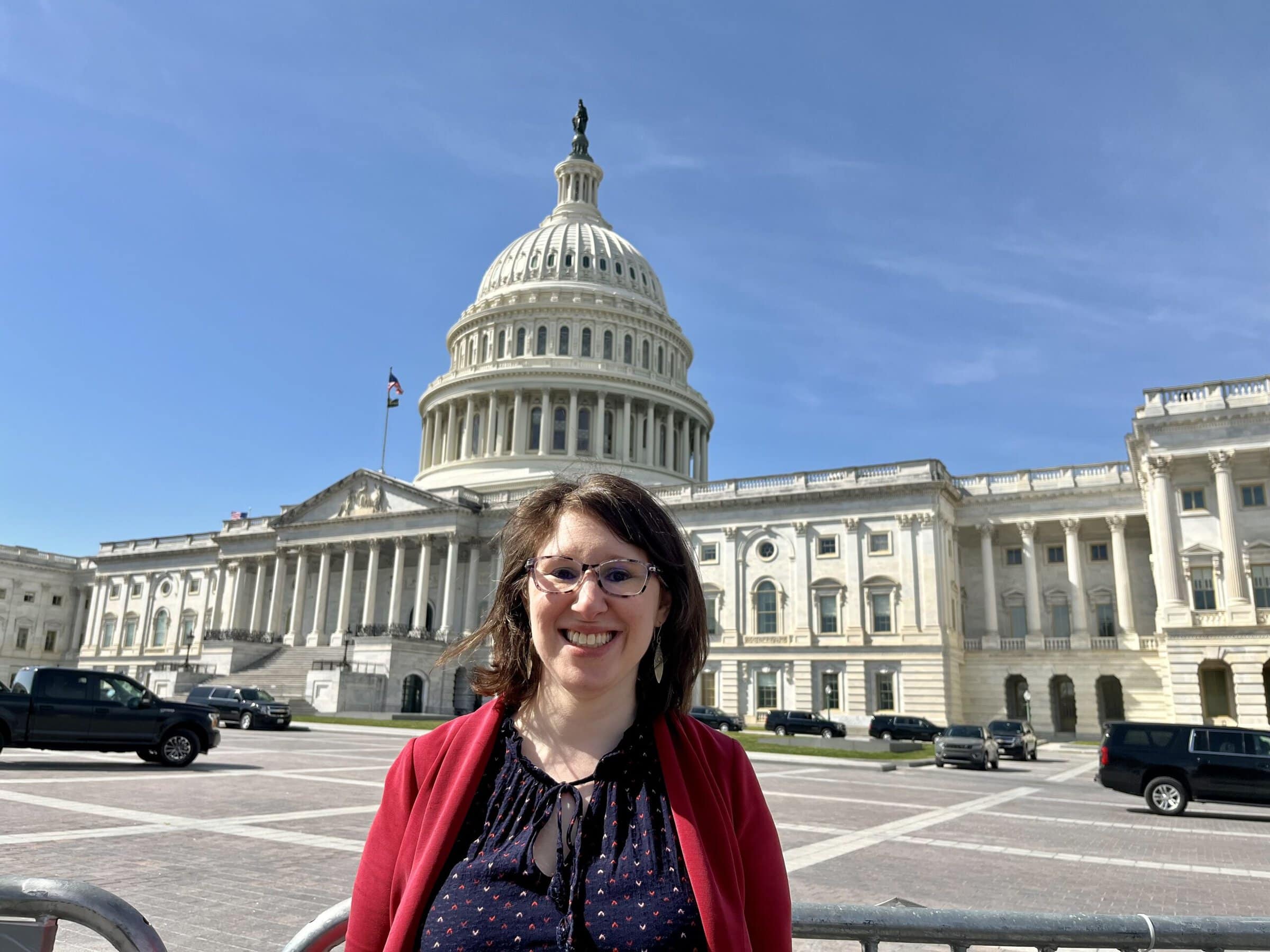 A brown-haired white woman, wearing a red cardigan and navy blue polka-dot blouse, standing in front of the United States Capitol building on a sunny day.