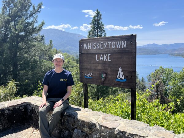 A woman in a ballcap, T-shirt, and long pants sits on a stone wall. Next to her is a wooden sign for Whiskeytown Lake. In the background are trees, mountains, and a lake.