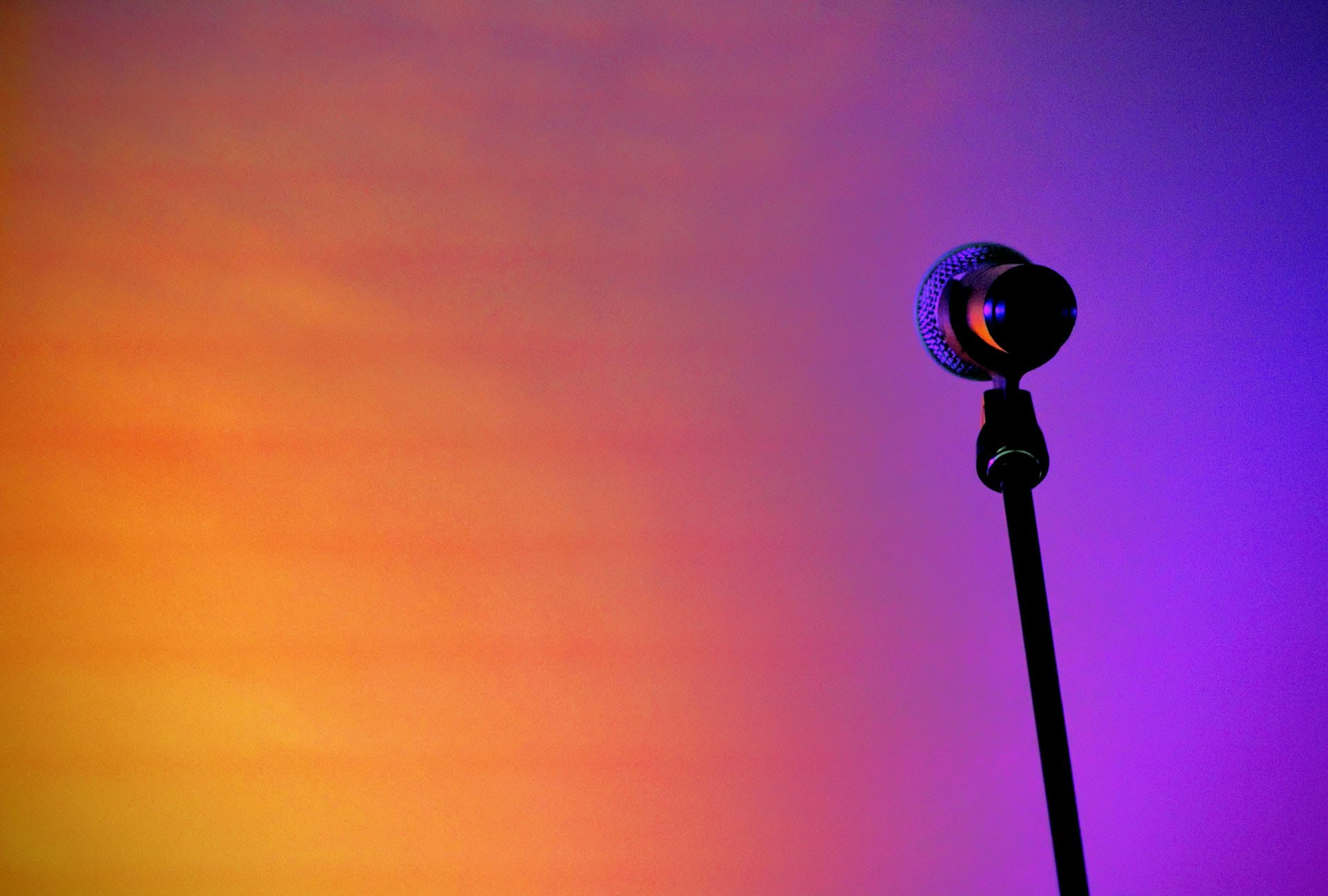 A three-quarter view of a black microphone and stand, in the right third of the image. The microphone is reflecting the yellow, orange, and purple gradient background.