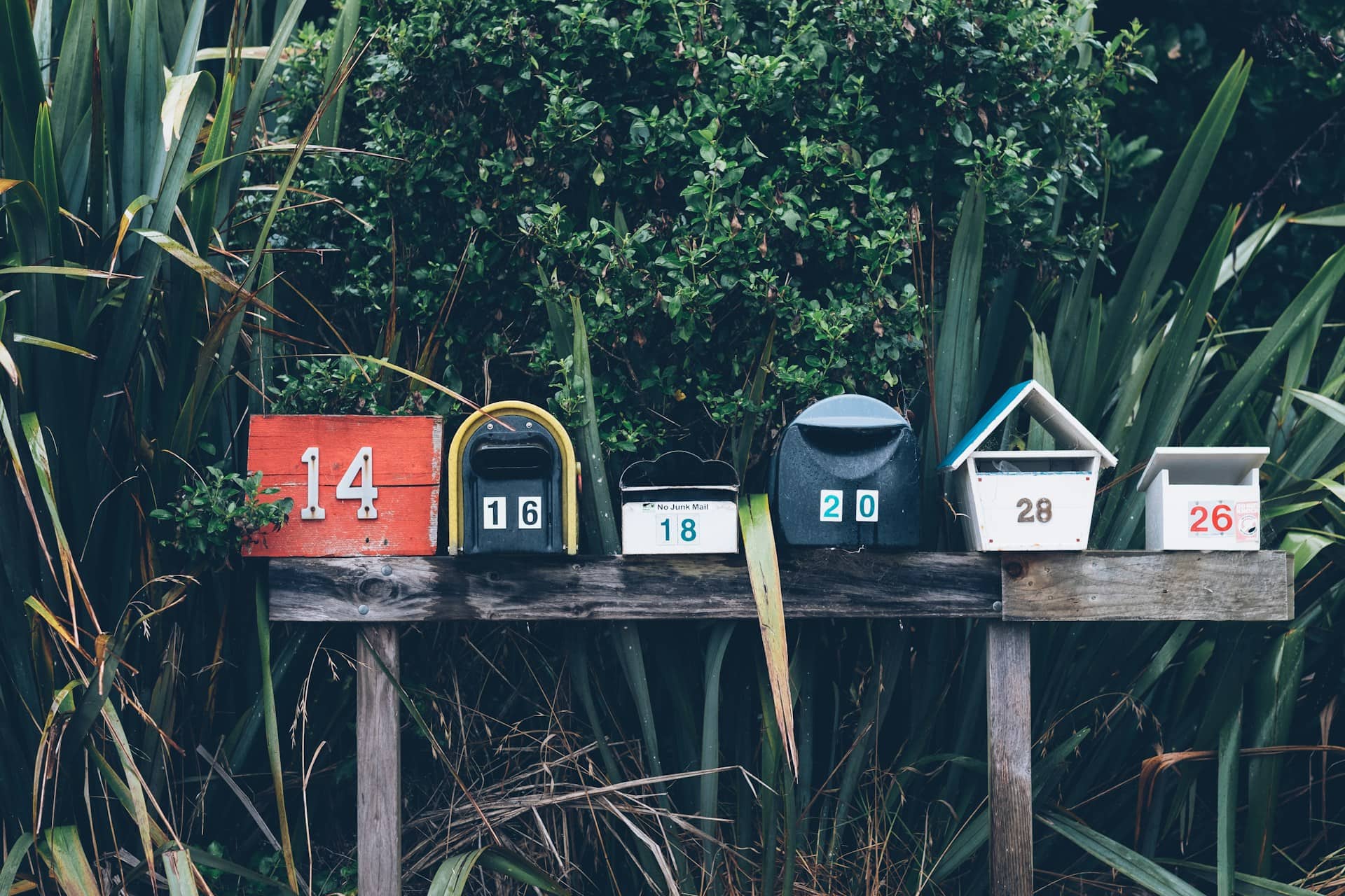 Series of differently sized and colored mailboxes resting on a weathered, wooden table against a background of dark green plants.