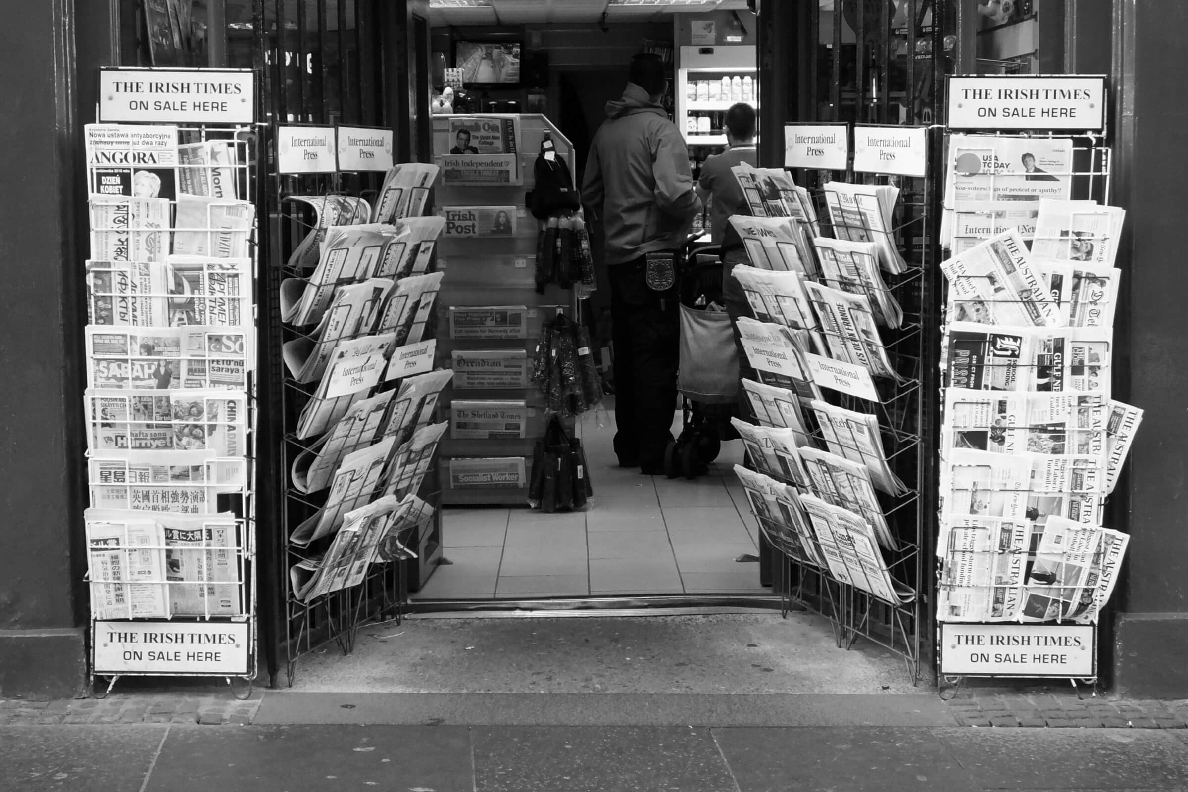 A black and white photo of a convenience store with newspaper display racks labeled "The Irish Times" and "International News" on either side of the open door.