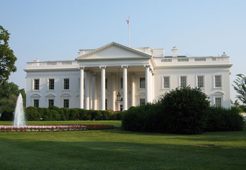 The United States White House, with a fountain and bush in front of it and blue skies behind.