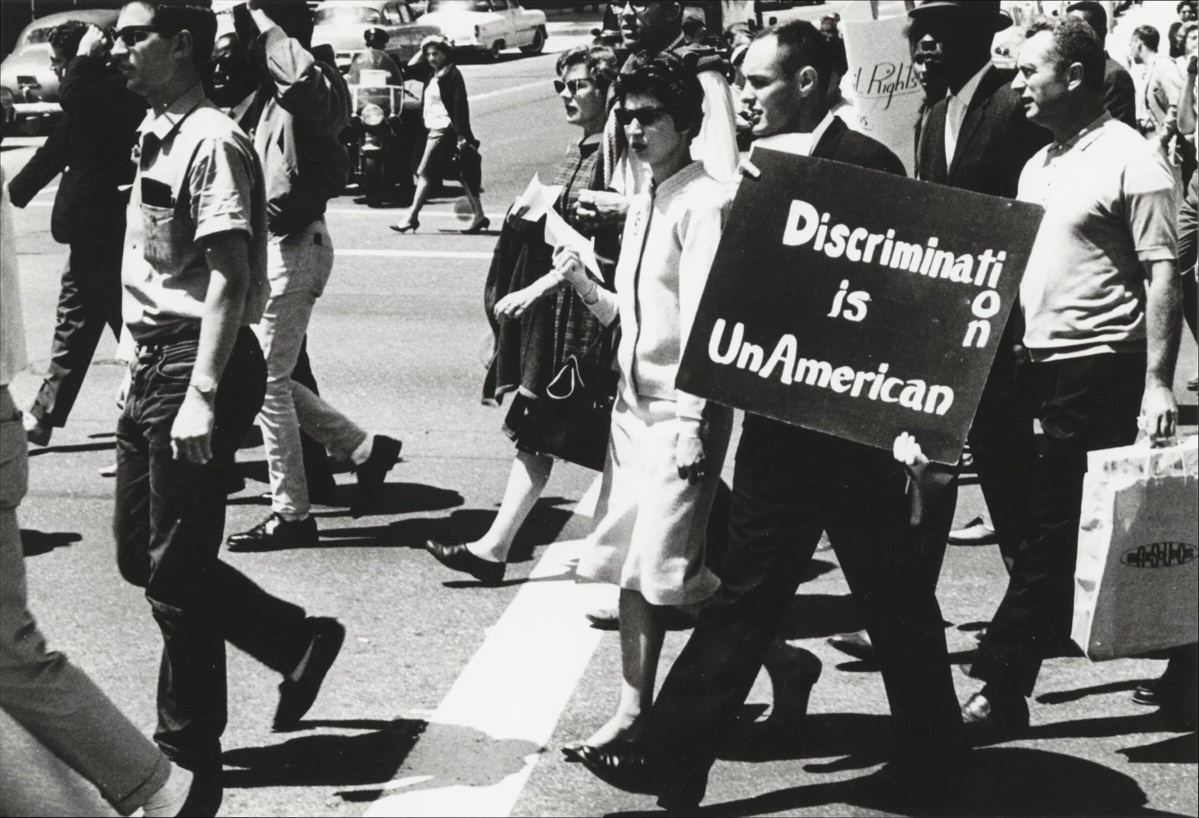 Black and white photograph depicting people marching together. One man holds a sign that reads "Discrimination is UnAmerican."