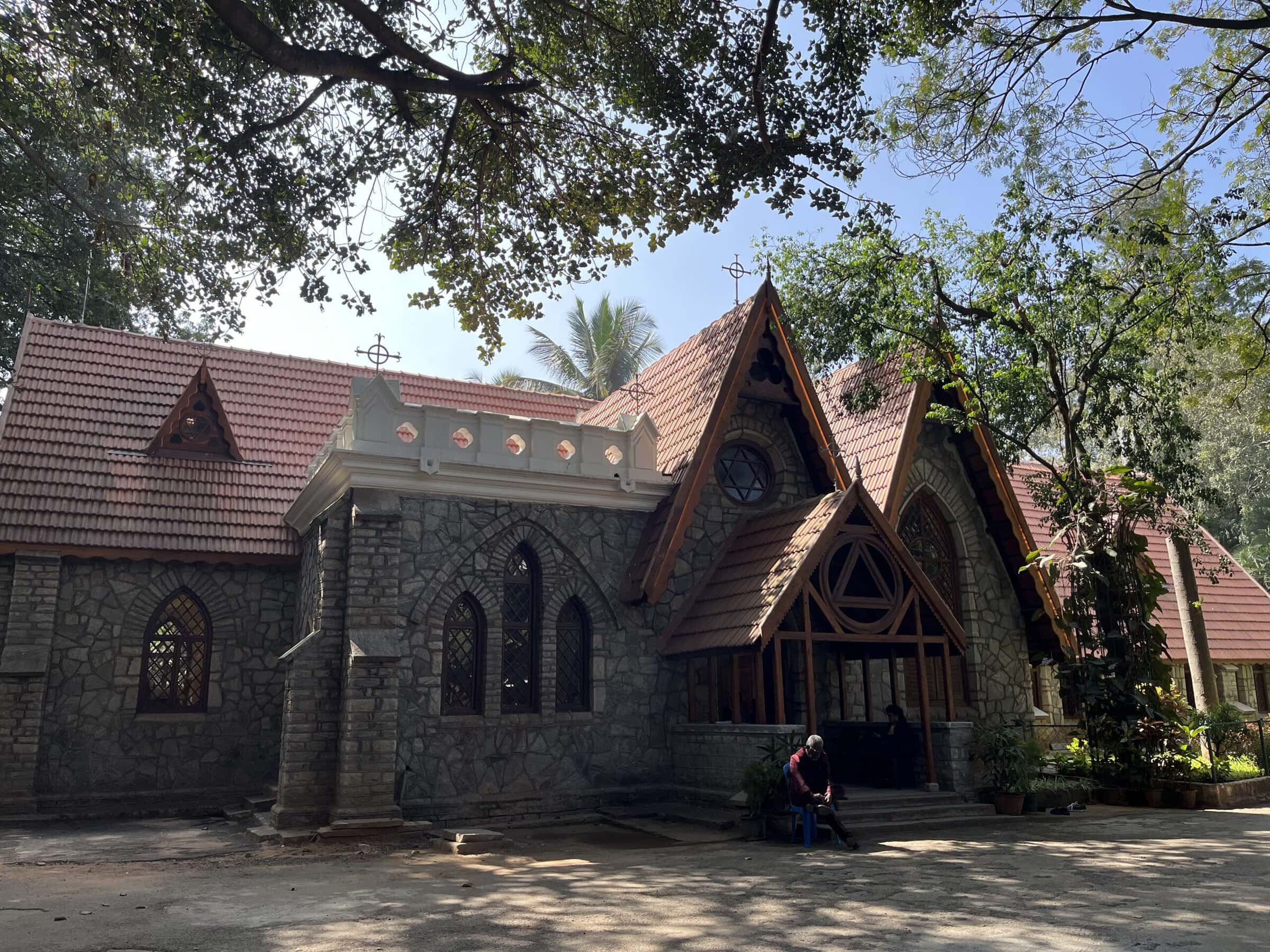 A stone church with red tile root, surrounded by trees. A man sits in a chair at the front of the church.