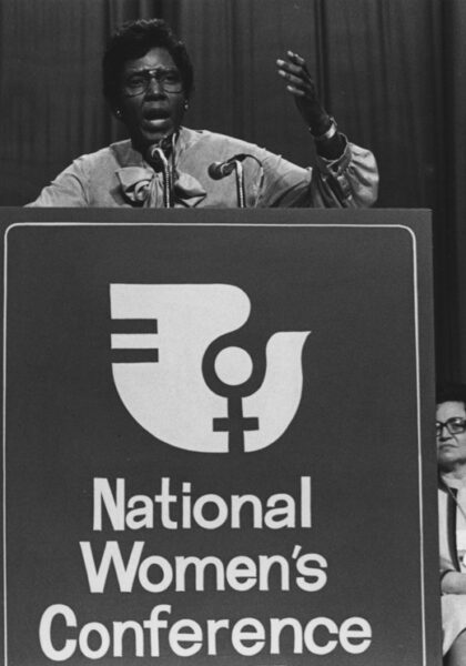 A black and white photo of a woman standing behind a podium, with the logo for the National Woman's Conference, speaking into a microphone