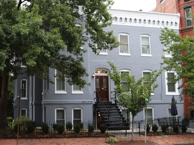 Gray-blue brick building with white trim and a black metal staircase in the front. Large tree is on the left, and brick sidewalk lines the front.