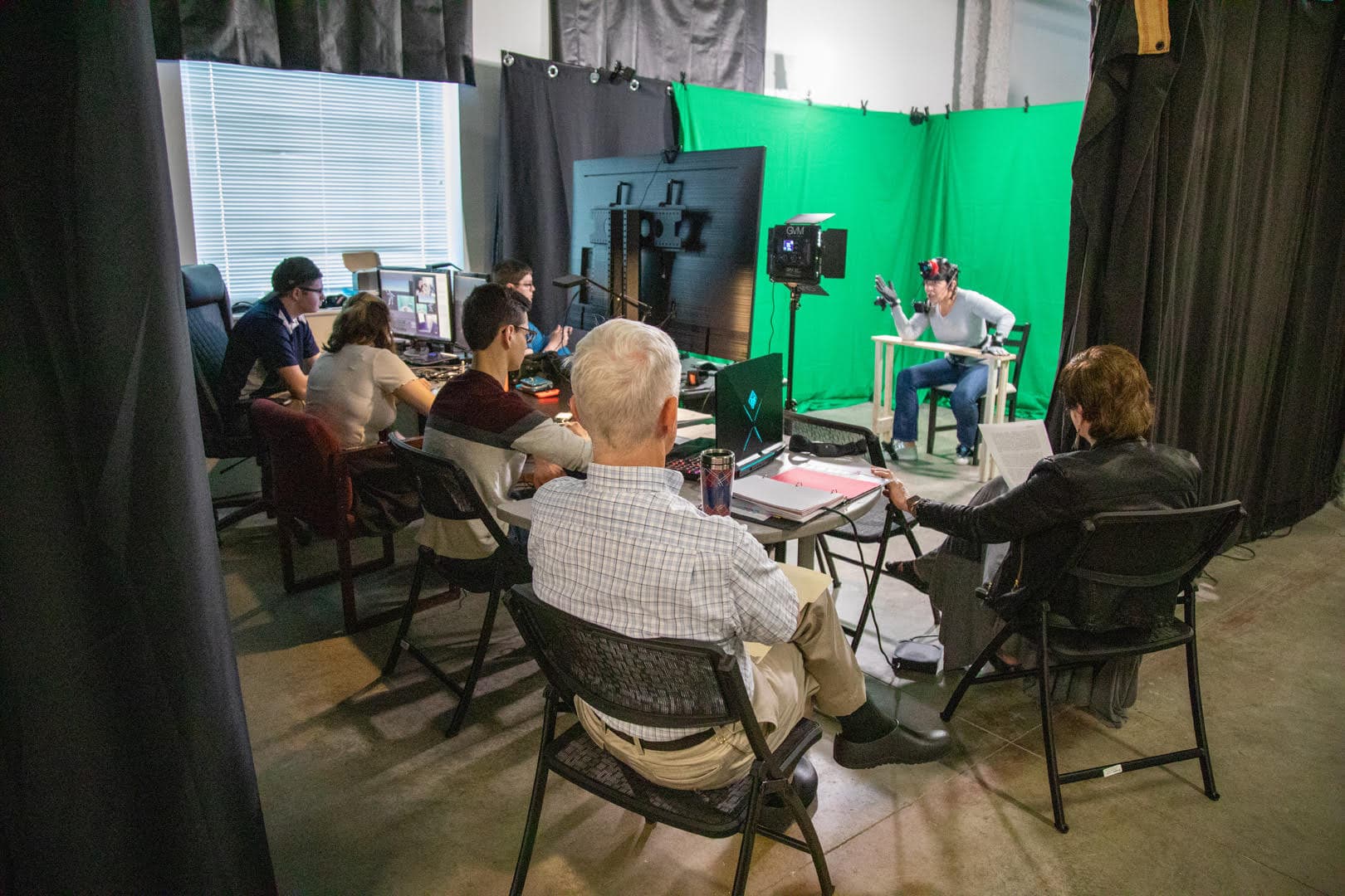 A group of people sitting around a table with laptops in front of them and a person posed sitting down in front of a green screen