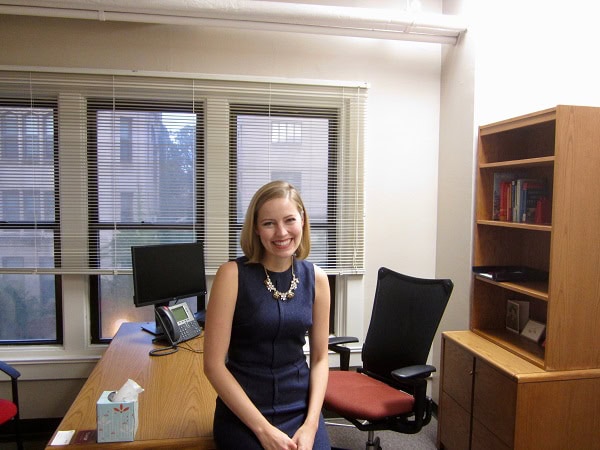 A women in a navy blue dress smiling while sitting on a desk.