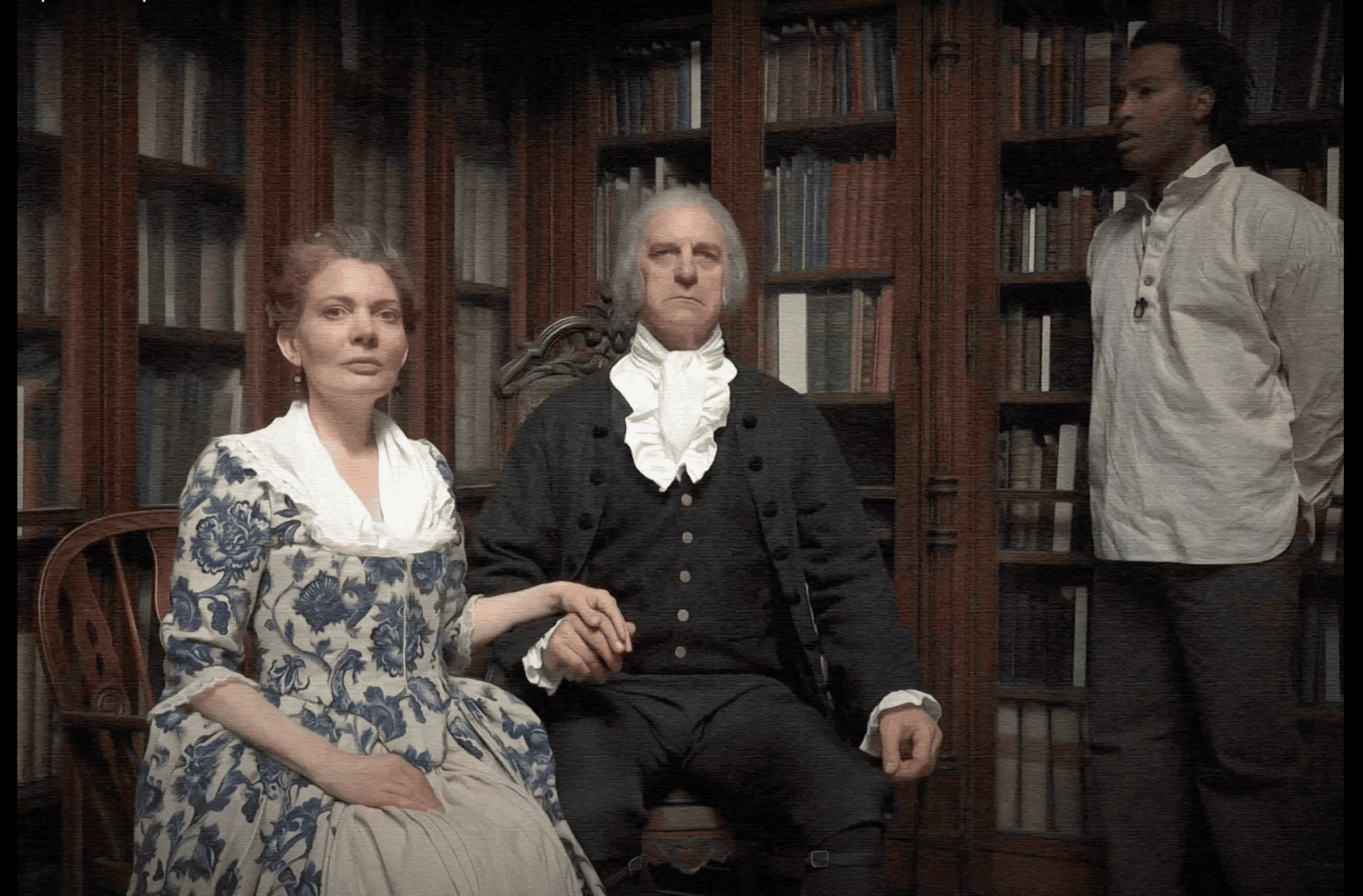 A white man and a white woman, well-dressed in an early American style, sit in front of elaborate wooden library shelving. A more simply dressed Black man stands to their left. The image is slightly distorted by horizontal lines, giving it the feel of an old television episode or a fuzzy memory.